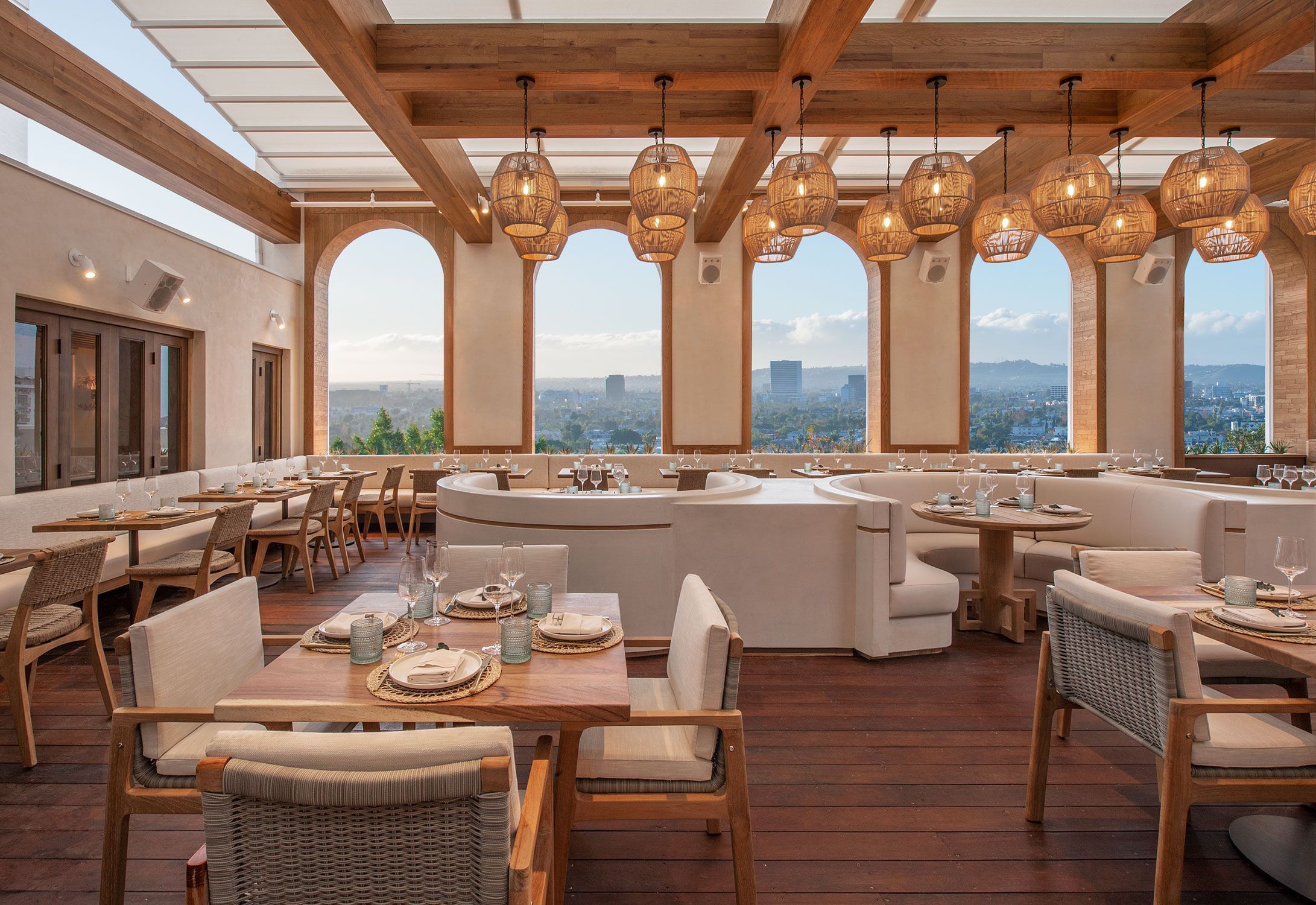 Dining area with wooden tables, beige cushioned chairs, large arch windows, and wicker pendant lights overlooking city views.