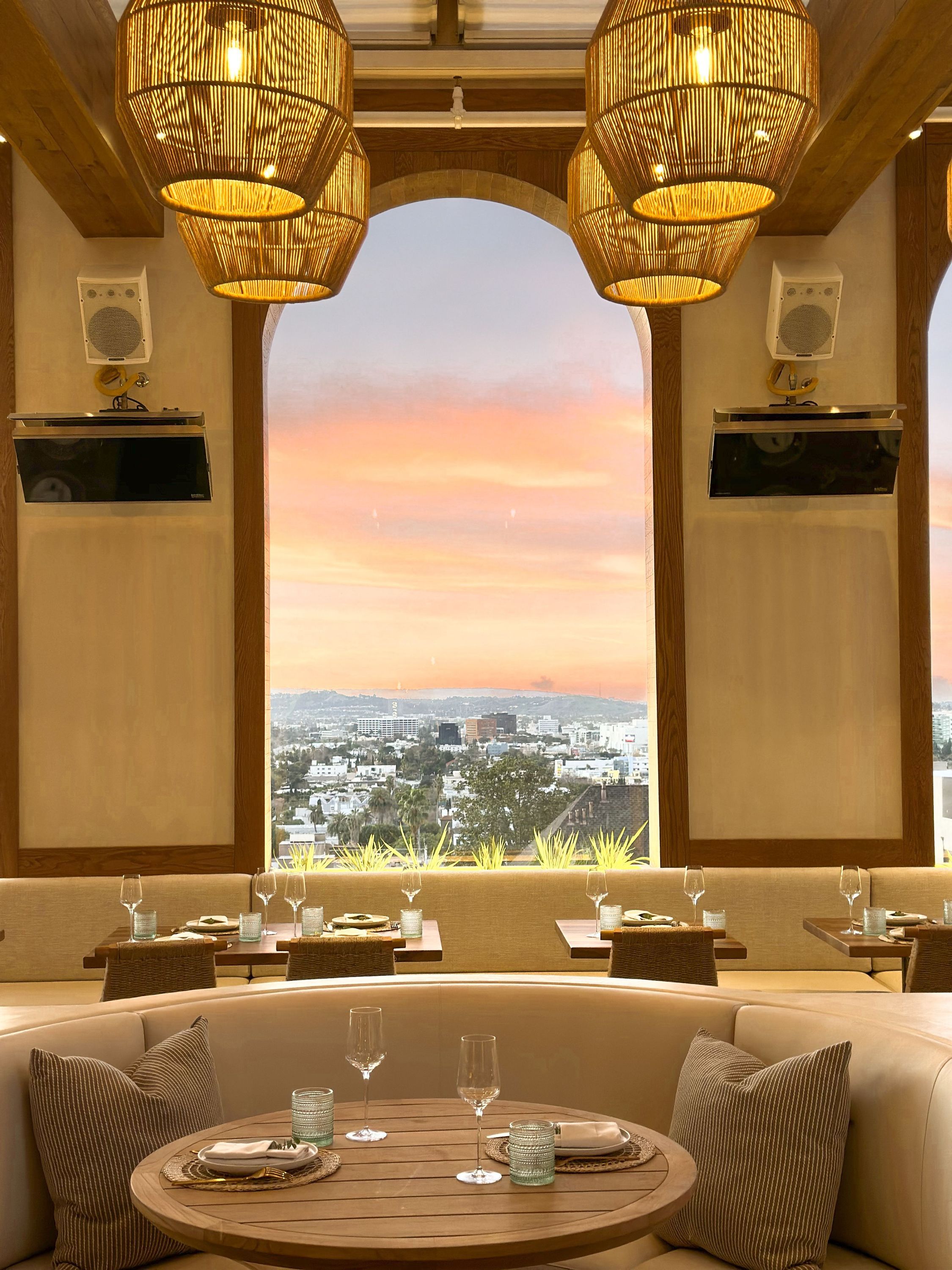 Dining area with round wooden table, cushioned seating, pendant lights, and panoramic city view through arched window.