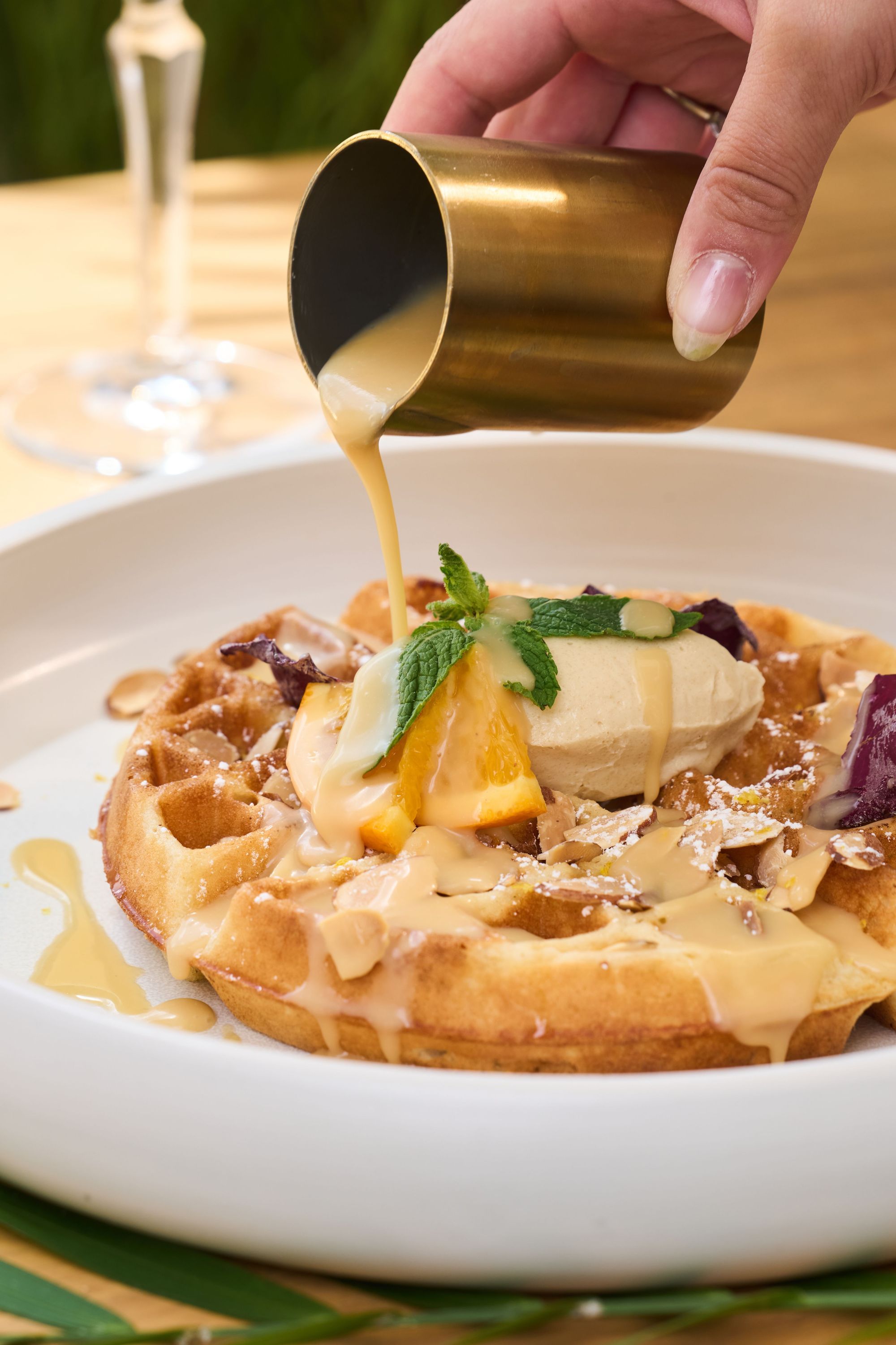 Waffle topped with cream, orange slice, and mint served with sauce poured from a small metal cup at brunch restaurant