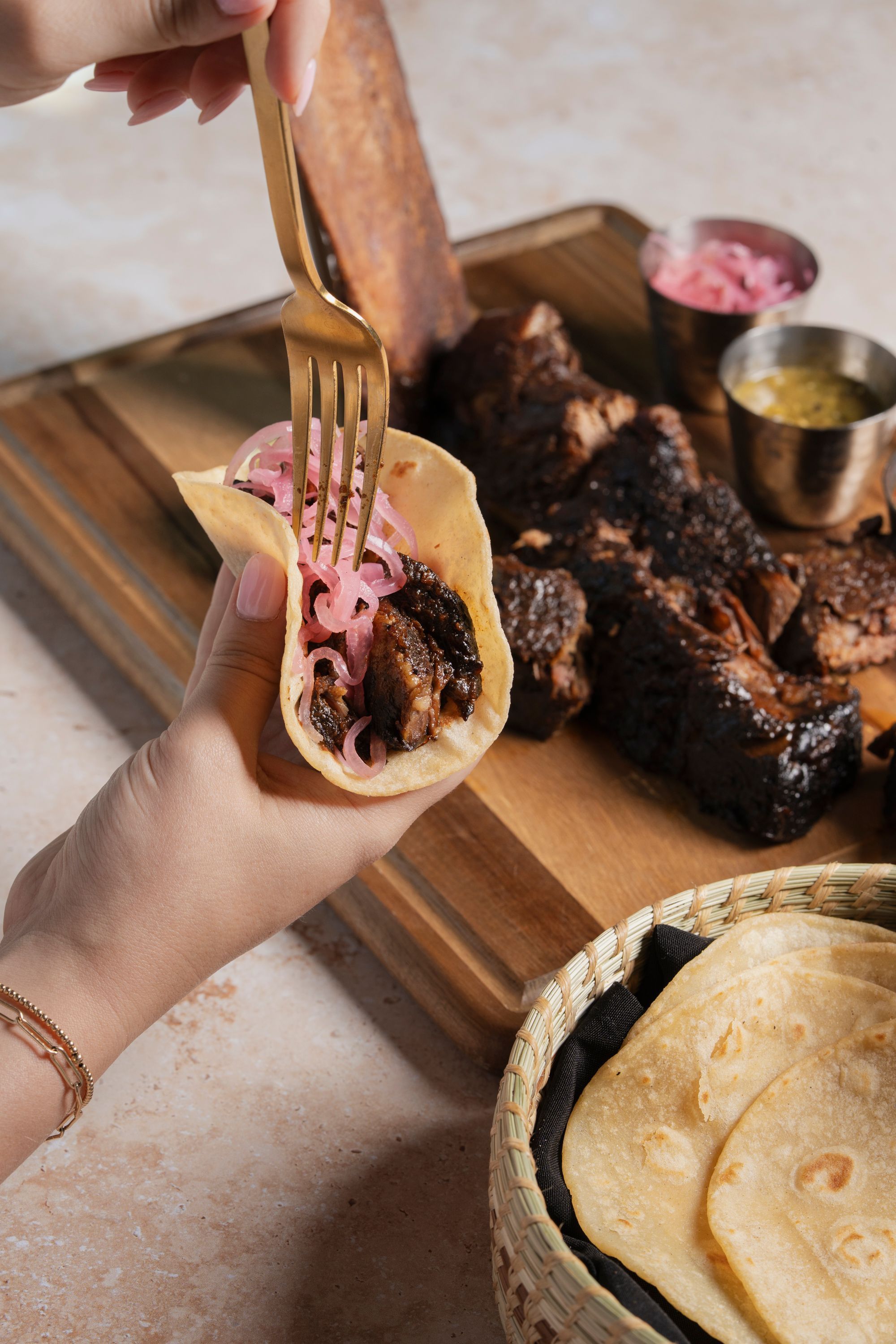 Grilled meat taco with pickled onions being prepared, accompanied by tortillas and grilled ribs on a wooden board.