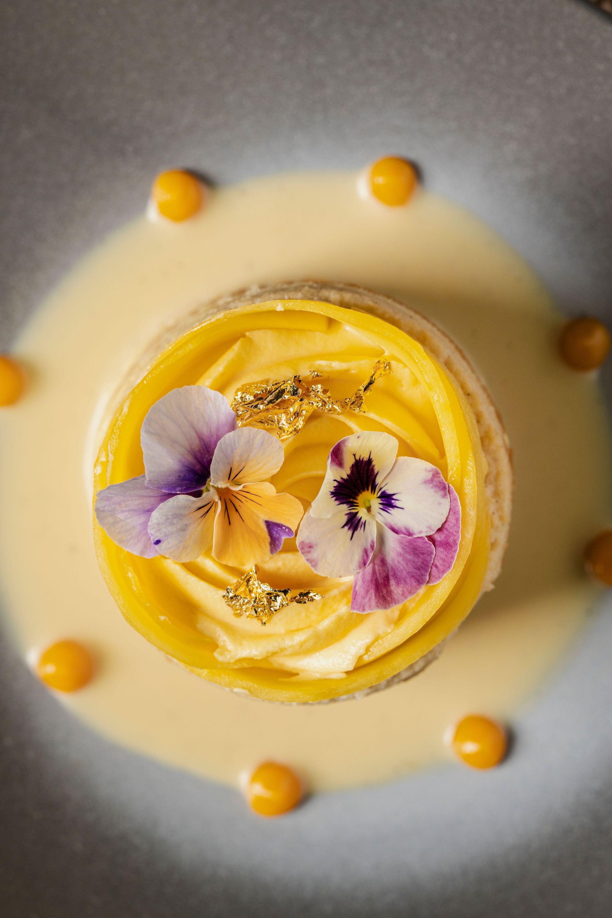 Delicate dessert with yellow cream, edible flowers, gold leaf accents, and small orange dots on a light sauce background.